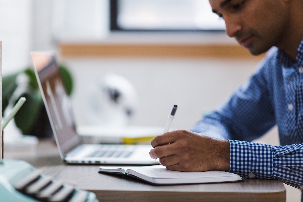 Man concentrates as he writes in a notepad next to his laptop